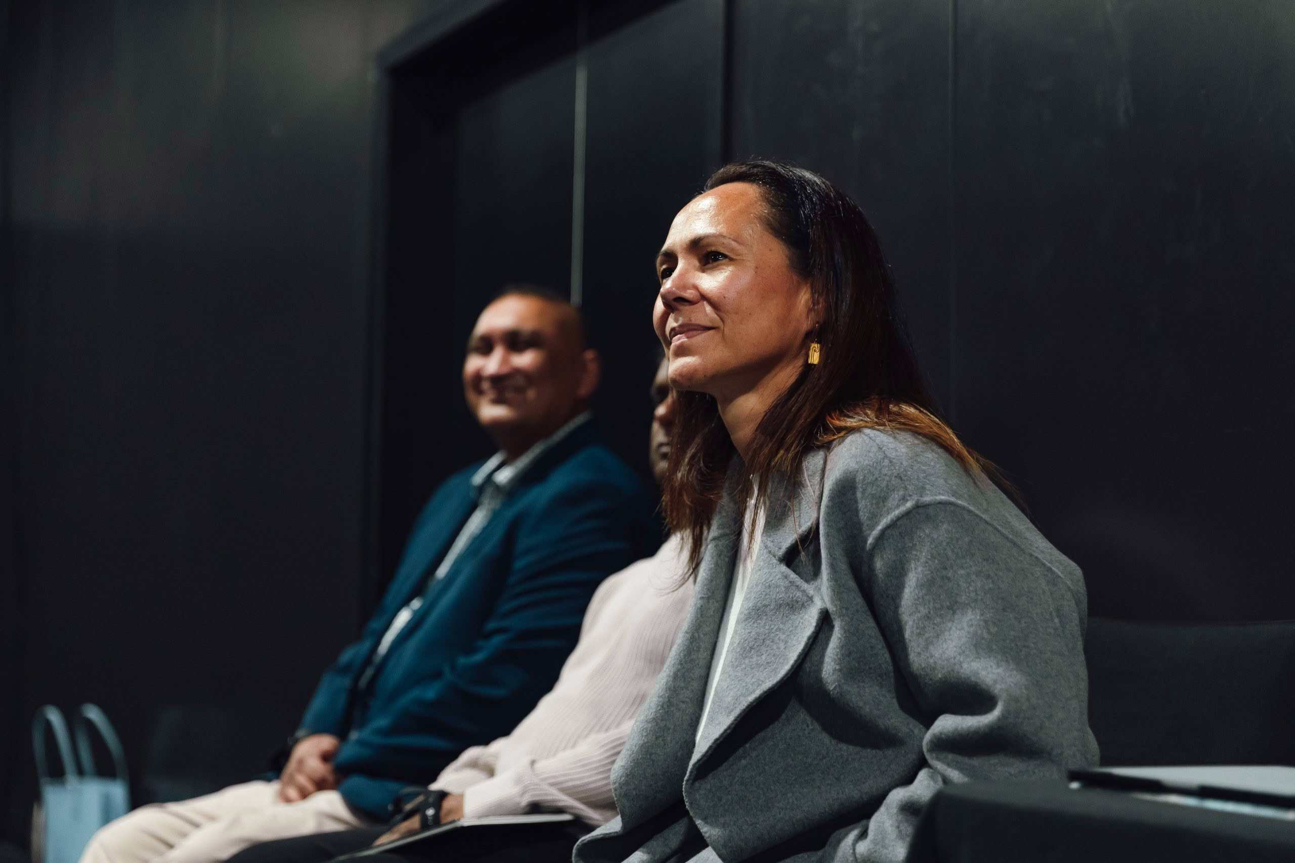 Melissa Wallace, a woman with long dark hair wearing a gray coat and gold earrings, smiling while seated at a professional event