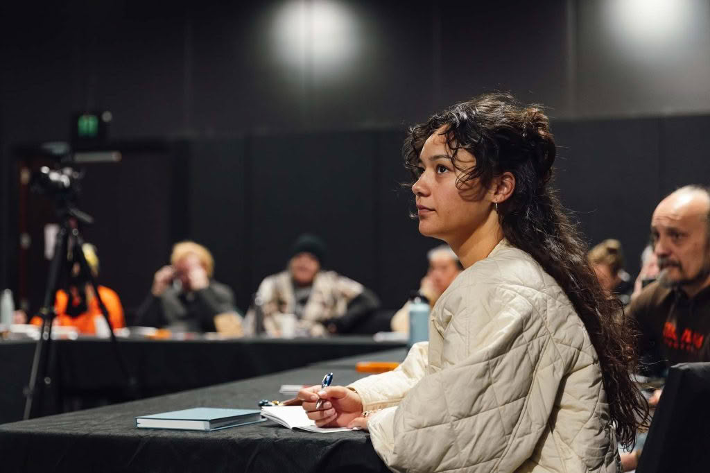 A young woman with curly dark hair wearing a cream quilted jacket, sitting at a conference table taking notes during a professional event.